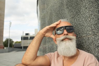 Hipster 'ı yakından gören olgun bir adam güneş gözlüğü takıyor ve şehrin ön cephesine yaslanıyor.