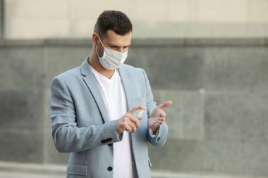 Şehirde yürüyen el dezenfektanı jeli kullanan tıbbi maske takan genç bir adam. Covid-19. Coronavirüs.