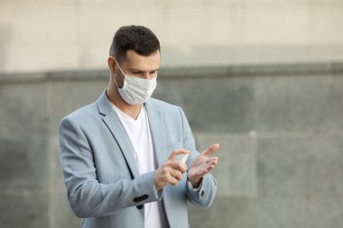 Şehirde yürüyen el dezenfektanı jeli kullanan tıbbi maske takan genç bir adam. Covid-19. Coronavirüs.