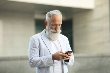 Sakallı kıdemli gülen iş adamı dışarıda duruyor ve cep telefonuyla kitap okuyor..