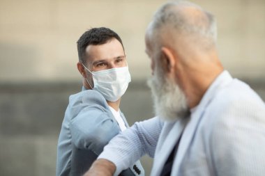 Koruyucu maskeler takan iki adam duvarın önünde selamlaşıyor - Kvid-19 koronavirüs salgınında yayılan virüs ve teması önlemek için yeni normal bir el sıkışma dirseği darbesi - sosyal uzaklık kavramı.