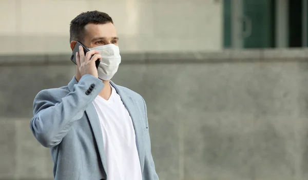 European young man in medical mask on his face calling by phone. Man in protective mask is protected from virus outdoor in street. Covid-19.