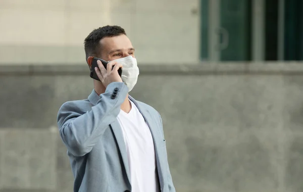European young man in medical mask on his face calling by phone. Man in protective mask is protected from virus outdoor in street. Covid-19.