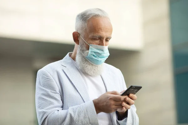 Bearded mature man with medical mask using mobile phone outdoor. Covid-19.