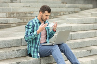 Adam dışarıda, merdivenlerde dizüstü bilgisayar üzerinde çalışırken koruyucu yüz maskesi takıyor..