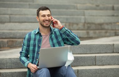 Genç serbest çalışan adam dışarıda bilgisayar laptopuyla çalışıyor. O, taş merdivenlerde oturuyor. Her yerde çalışma kavramı