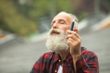 Sakallı yaşlı adam duman üflüyor. Elektronik sigara içiyorum. Stres atma konsepti. Sigara içme cihazı. Uzun sakallı adam sigara içmekten rahatlamış. Sakallı adam buharlaşıyor. IQOS.