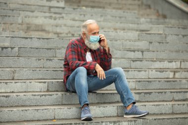 Yaşlı insanlar için en önemli koruyucu maske. Dışarıda telefon kullanan yaşlı sakallı bir adam. Covid 19.