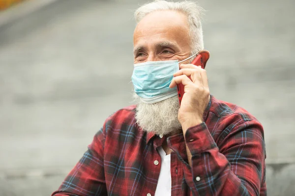 Protective mask most important for old people. Bearded senior man using phone outdoor. Covid-19.