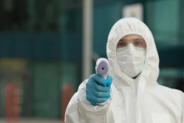 Male doctor use infrared thermometer to check body temperature for virus Covid-19 symptoms with the isolation gown or protective costume and surgical face masks outside on street.