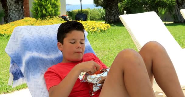 サンベッドの上に敷設 ポテトチップスを食べるかわいいプレティーン少年の肖像画 — ストック動画