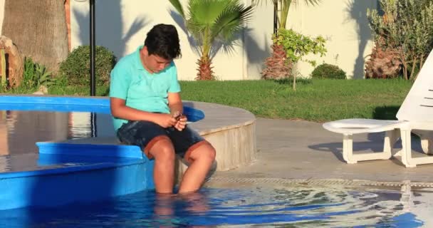 スイミング プールの端に座っていると 日当たりの良い夏の日にスマート フォンを使用してかわいい 代の少年の肖像画 — ストック動画