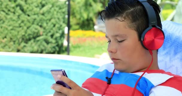 一个可爱的十几岁男孩的肖像坐在日光浴附近的游泳池和使用智能手机在一个阳光明媚的夏日 互联网通信和人的概念 智能手机成瘾 — 图库视频影像