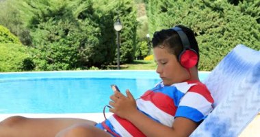 Yüzme havuz kenarında şezlong üzerinde oturan ve bir güneşli yaz gününde smartphone kullanarak kulaklık ile sevimli bir genç çocuk portresi. Teknoloji, Internet iletişim ve insan kavramı, smartphone bağımlılığı
