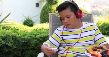 Kulaklıklı, müzik dinleyen ve yeşil bahçede farklı meyveler yiyen mutlu bir okul çocuğunun portresi.