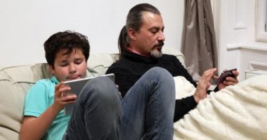 Dijital tablet ve evde kanepede oturan akıllı telefon ile beyaz bir adam ve çocuk portresi. Akıllı telefon bağımlılığı