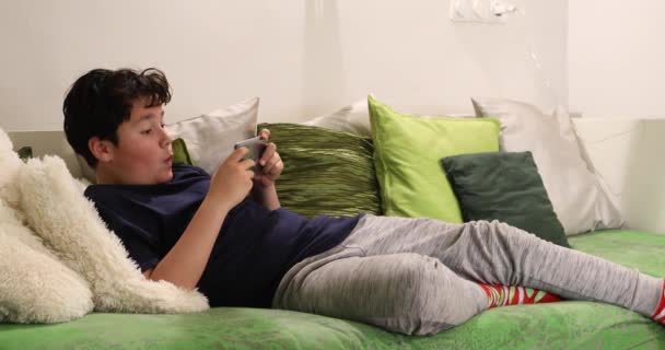 自宅でソファーに座っているスマートフォンで預け少年の肖像 子供は タイピング ゲームをプレイするために見ています インターネット通信と人の概念 スマートフォン中毒 — ストック動画
