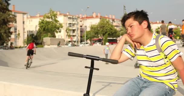 英俊的不幸的青春期男孩与滑板车独自坐在滑板公园 — 图库视频影像