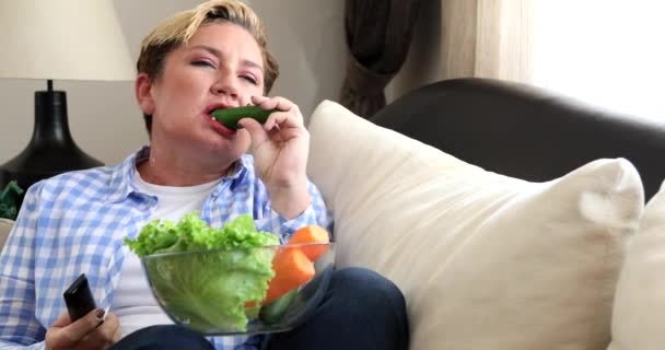 Retrato de una mujer comiendo alimentos frescos y viendo la televisión en casa — Vídeo de stock