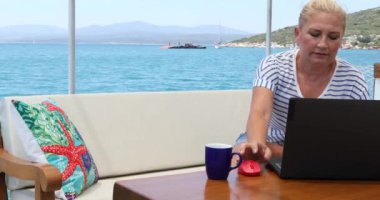 Orta yaşlı serbest çalışan bir kadının portresi yaz tatilinde bir yatın güvertesinde dizüstü bilgisayarla oturuyor.