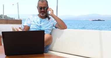 Serbest çalışan bir iş adamının portresi bir yatın güvertesinde dizüstü bilgisayarı ve akıllı telefonuyla yaz tatilinde oturuyor.