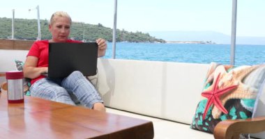 Orta yaşlı serbest çalışan bir kadının portresi yaz tatilinde bir yatın güvertesinde dizüstü bilgisayarla oturuyor.