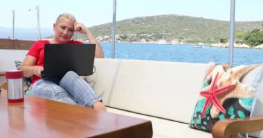 Orta yaşlı serbest çalışan bir kadının portresi yaz tatilinde bir yatın güvertesinde dizüstü bilgisayarla oturuyor.