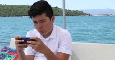 Beyaz tenli genç yaz tatilinde yatta akıllı telefonla video oyunu oynuyor.