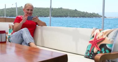 Orta yaşlı güzel bir kadının portresi yaz tatilinde bir yatın güvertesinde elinde akıllı bir telefonla oturuyor.