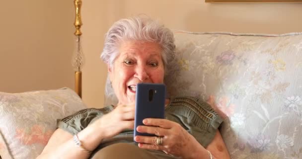 一个快乐的高加索老年妇女坐在沙发上 家里手机看起来很震惊的画像 — 图库视频影像