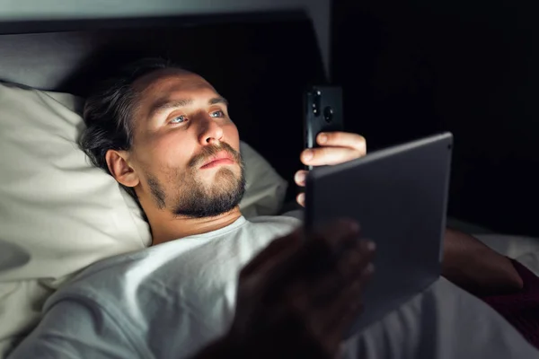 Pemuda Berjanggut Berbaring Tempat Tidur Sambil Melihat Telepon Dan Tablet — Stok Foto