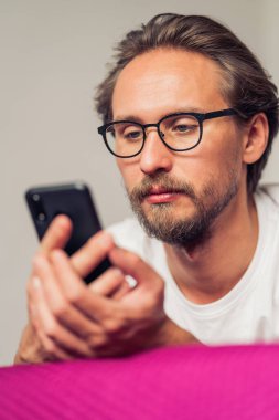 Yatakta yatarken gözlük takan oldukça ciddi bir genç adam.