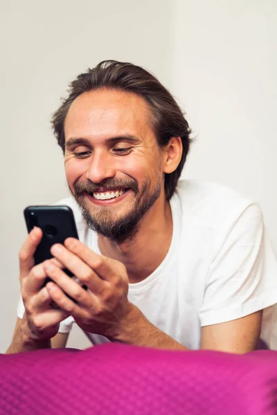 Beau jeune homme souriant couché sur le lit tout en tenant son mobile Image En Vente