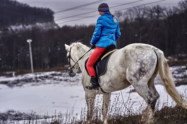 Young woman rider in a blue blazer and sporting a cap for a walk on a white horse on a cloudy winter day.