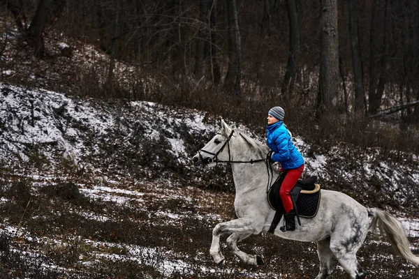 Young woman rider in a blue blazer and sporting a cap for a walk on a white horse on a cloudy winter day.