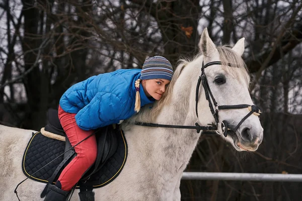 Young woman rider in a blue blazer and sporting a cap for a walk on a white horse on a cloudy winter day.