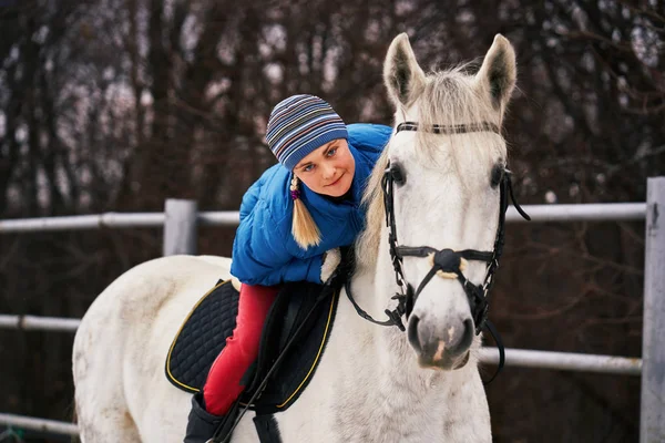 Young woman rider in a blue blazer and sporting a cap for a walk on a white horse on a cloudy winter day.