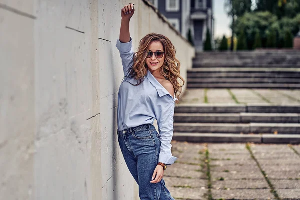 Young happy woman with long brown hair in sunglasses, blue jeans, blue shirt walks through the old town on a cloudy summer day.