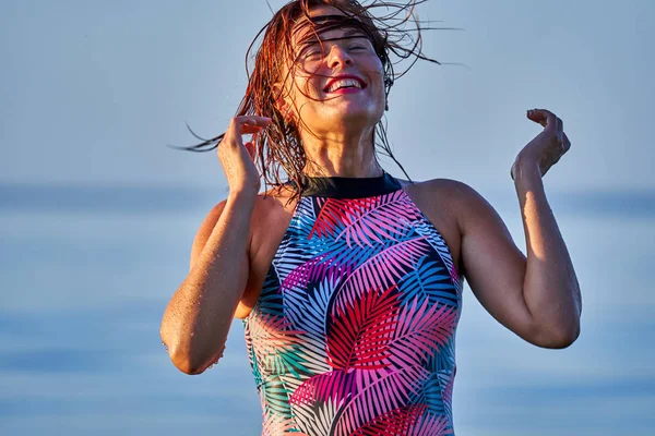 Portrait of a red-haired wet laughing woman of middle age in a swimsuit on a summer evening in the light of the setting sun. Close-up.