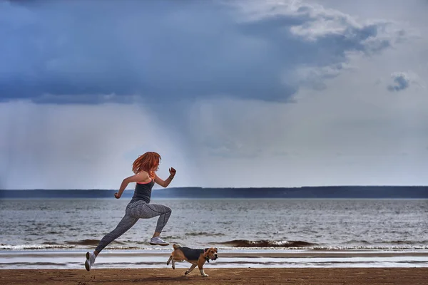 A red-haired middle-aged woman in sportswear runs along the sandy shore of a large river with her puppy on a cloudy summer morning. Healthy lifestyle.