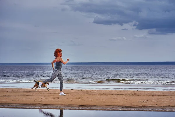 A red-haired middle-aged woman in sportswear runs along the sandy shore of a large river with her puppy on a cloudy summer morning. Healthy lifestyle.