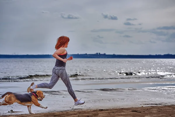 A red-haired middle-aged woman in sportswear runs along the sandy shore of a large river with her puppy on a cloudy summer morning. Healthy lifestyle.