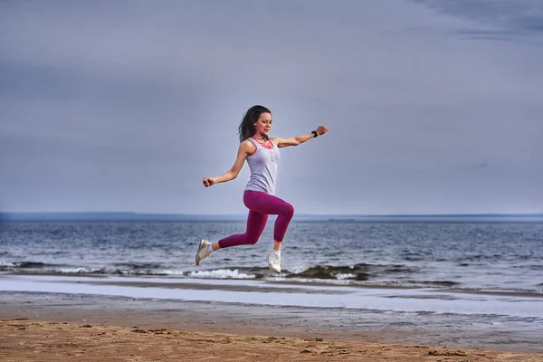 Young woman with black hair jumps while jogging. A woman is engaged in gymnastics in the spring morning on the sandy bank of a large river. Cloudy morning.