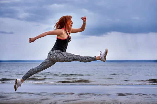 A middle aged woman with red hair jumps while jogging. A woman practices gymnastics on the sandy bank of a large river. Cloudy spring morning.