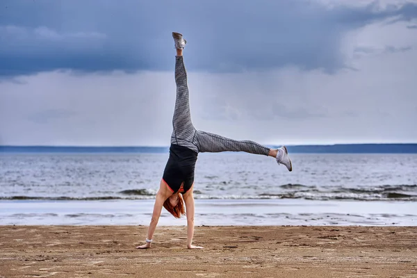 A middle-aged woman with red hair does gymnastics on the sandy shore of a large river. Cloudy spring morning.