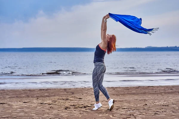 Slim red-haired middle-aged woman in sports form is engaged in dancing with a blue scarf. A woman is engaged on the sandy shore of a large river. Cloudy summer morning.
