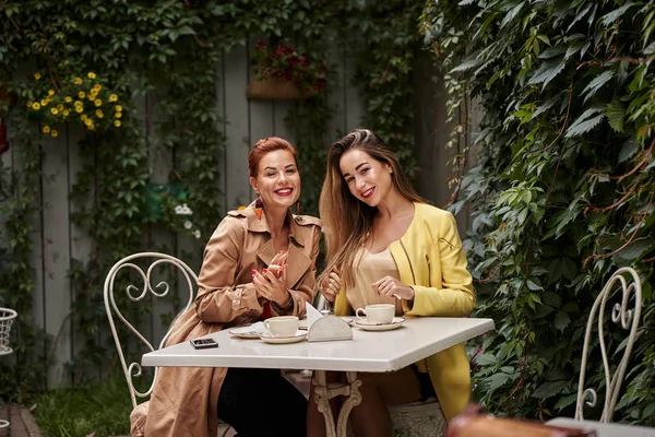 A middle-aged red-haired woman in a brown coat and a brown-haired young woman in a yellow coat are sitting in a summer street cafe and  talking to each other.