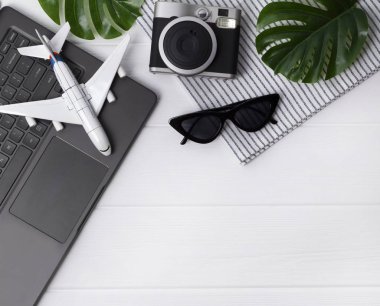 Seyahat flatlay: dizüstü bilgisayar, kamera, güneş gözlüğü, monstera yaprakları, uçak ve havlu, kopya alanı ile ahşap masa üzerinde.