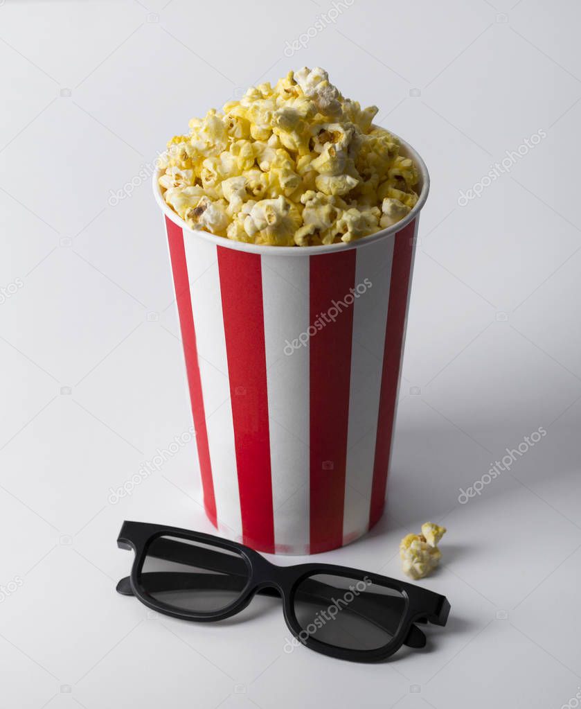 Popcorn in striped red bucket with 3d glasses at white table.