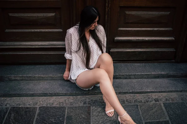 欧洲年轻性感漂亮的女孩短裤和白色衬衫坐在房子下的长腿在混凝土俯瞰门 等待体贴的概念 — 图库照片